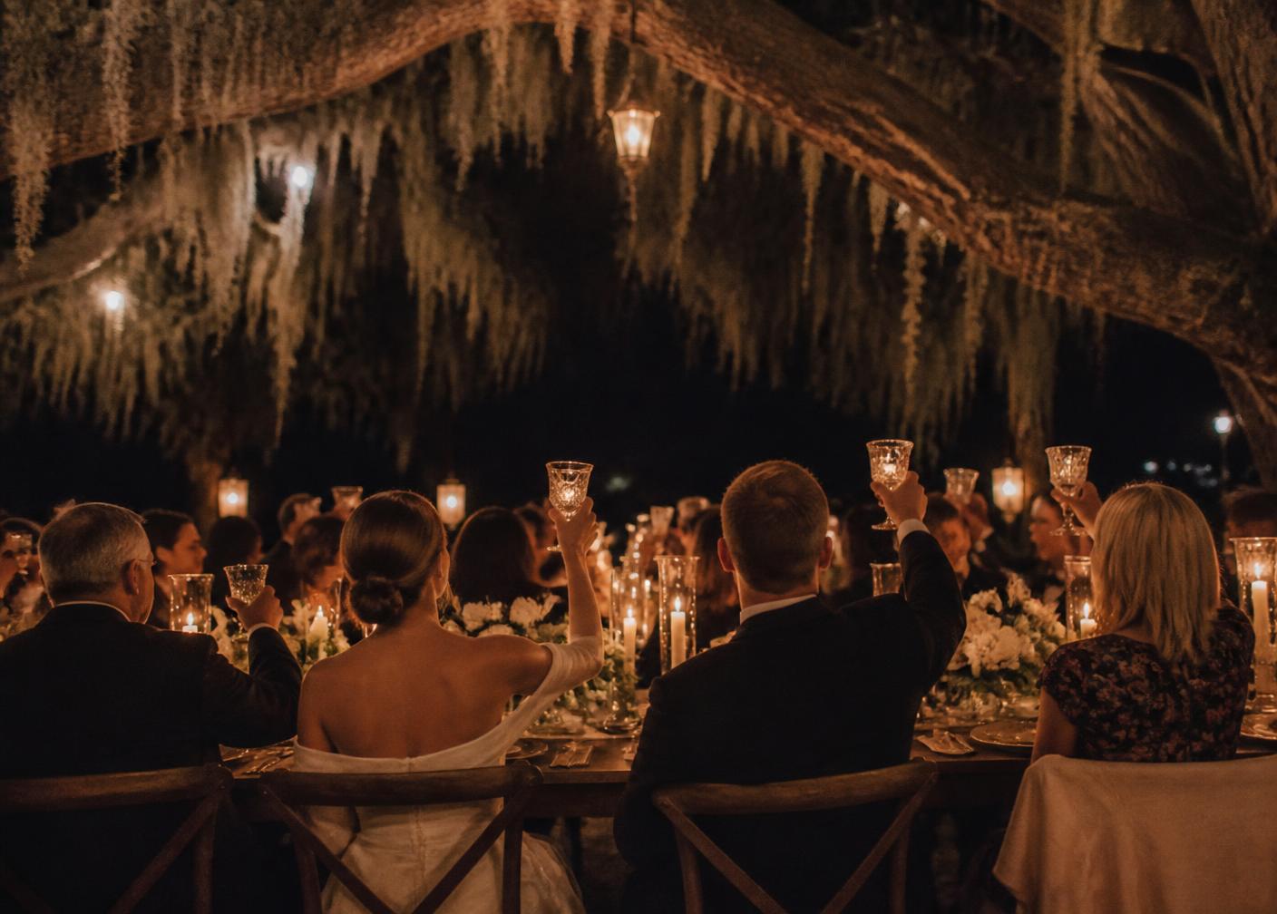 Guests raising glasses at a candlelit night-time table under Spanish moss